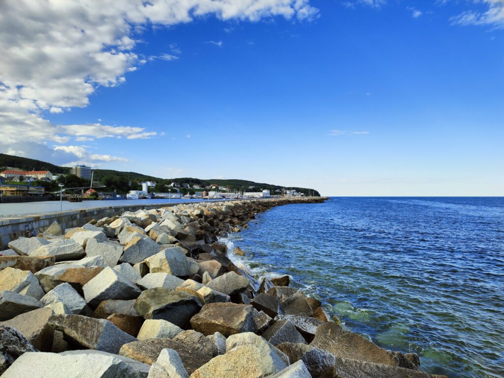 Große Felsbrocken schützen die Mole einer Küstenstadt; links Gebäude, rechts weites blaues Meer unter lockeren Wolken