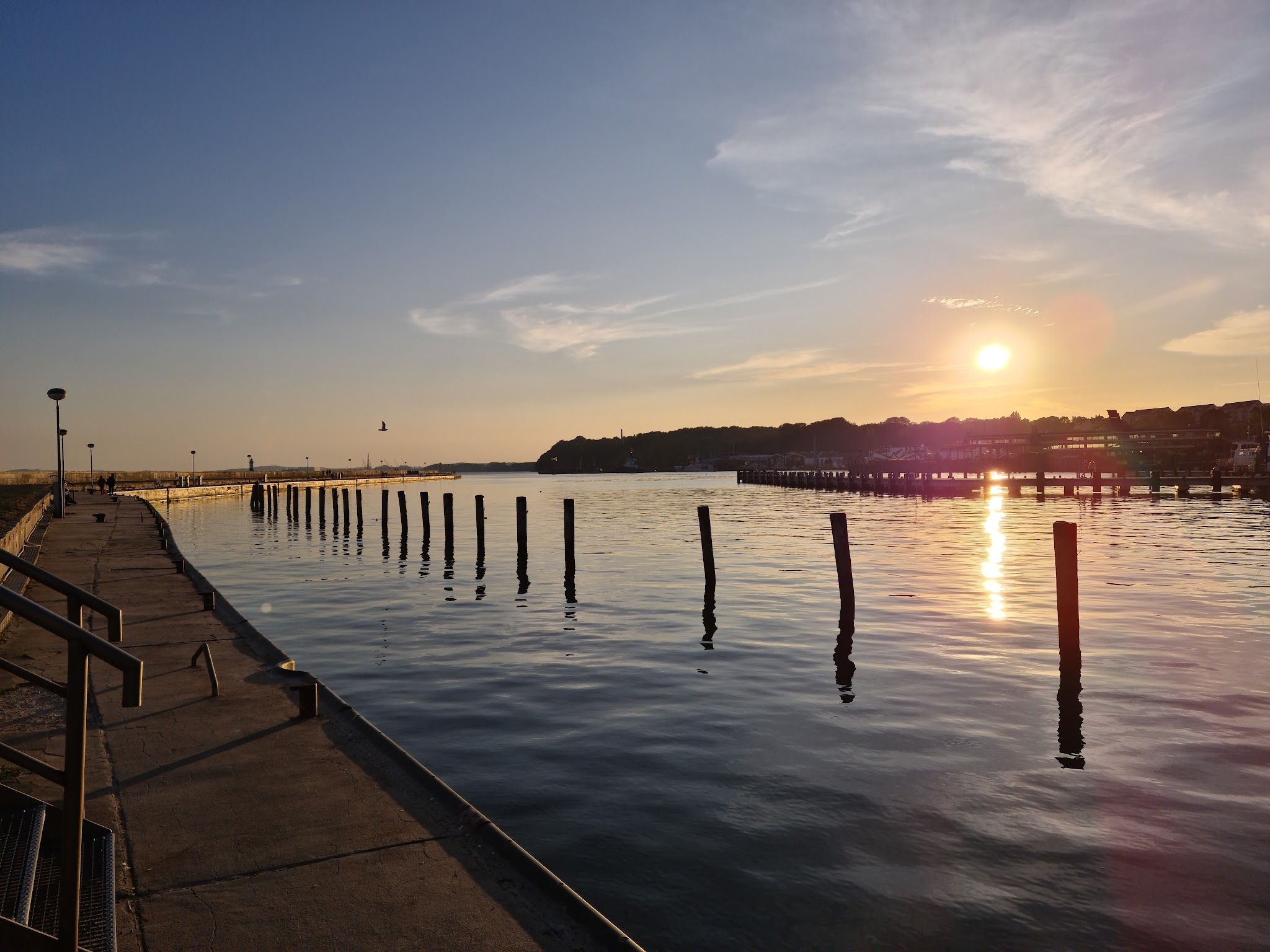 Sonnenuntergang über ruhigem Hafen; Stegpfähle spiegeln sich im Wasser, Promenade links führt zum Horizont.