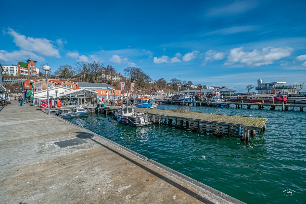 Kleiner Hafen mit bunten Fischerbooten, Holzstegen und orangefarbenem Gebäude unter blauem Himmel.