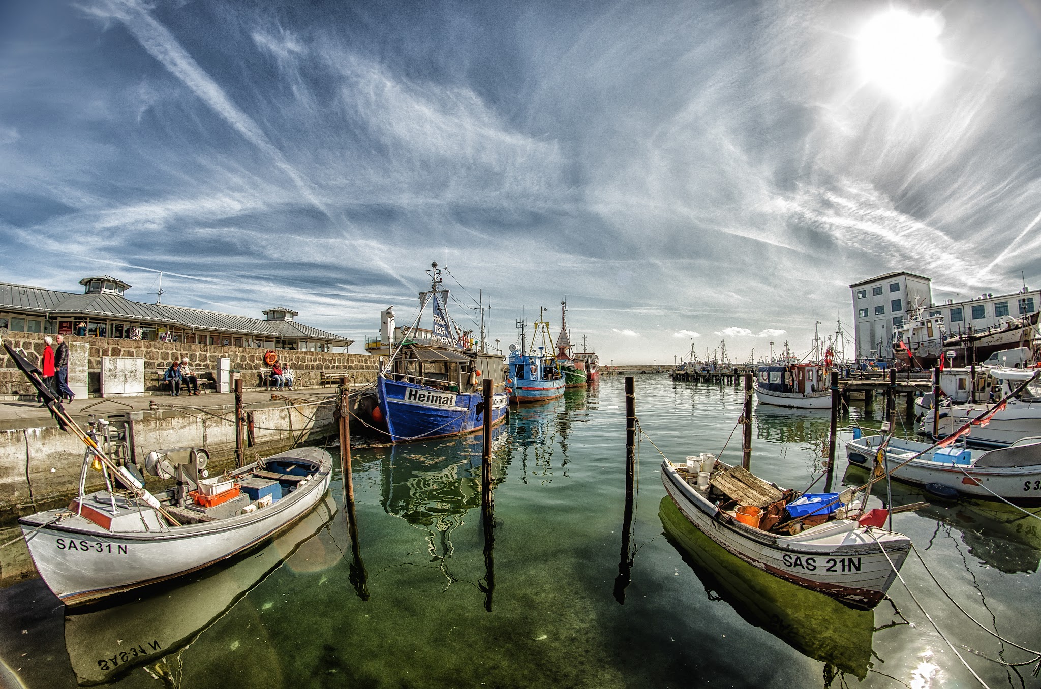 Kleiner Fischerhafen: Boote am Kai, klares Wasser, strahlende Sonne und Wolkenschleier am weiten Himmel.