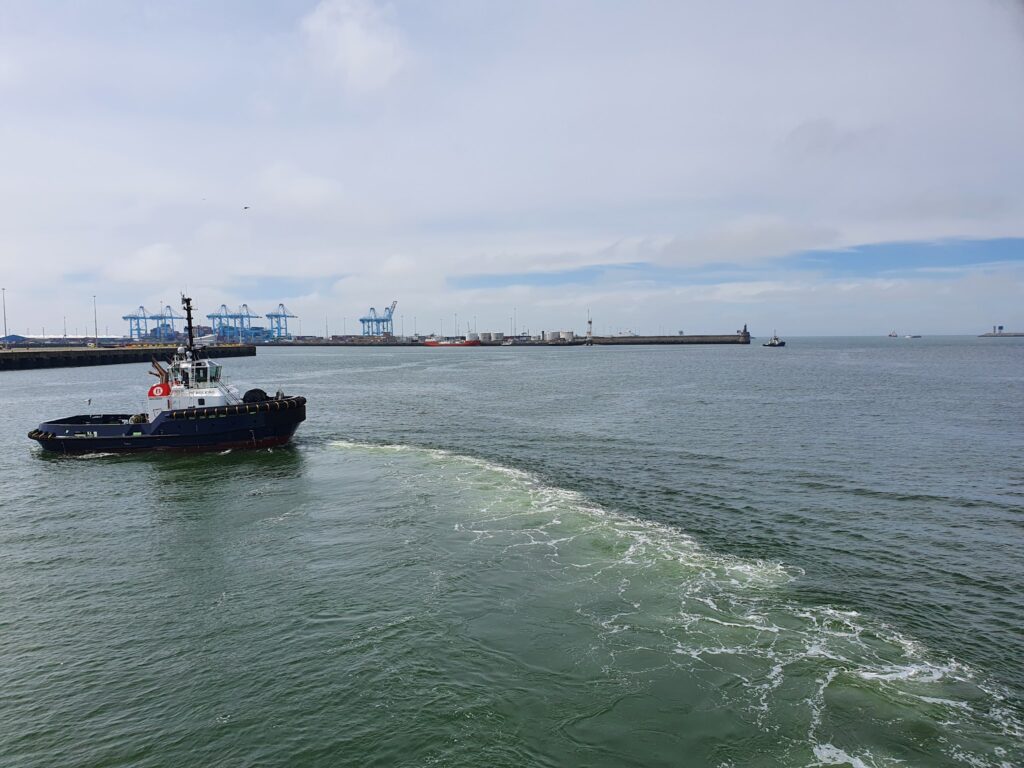 Schlepper dreht im Hafen, hinter ihm Wellenwirbel; am Kai blaue Containerkräne unter teils bewölktem Himmel.