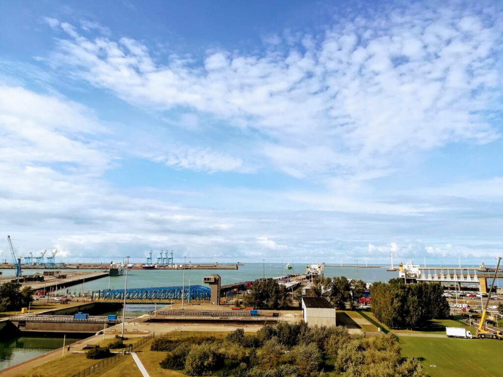 Hafenanlage mit blauen Kränen und Piers an türkisfarbener See unter locker bewölktem Himmel; Grünflächen vorne.