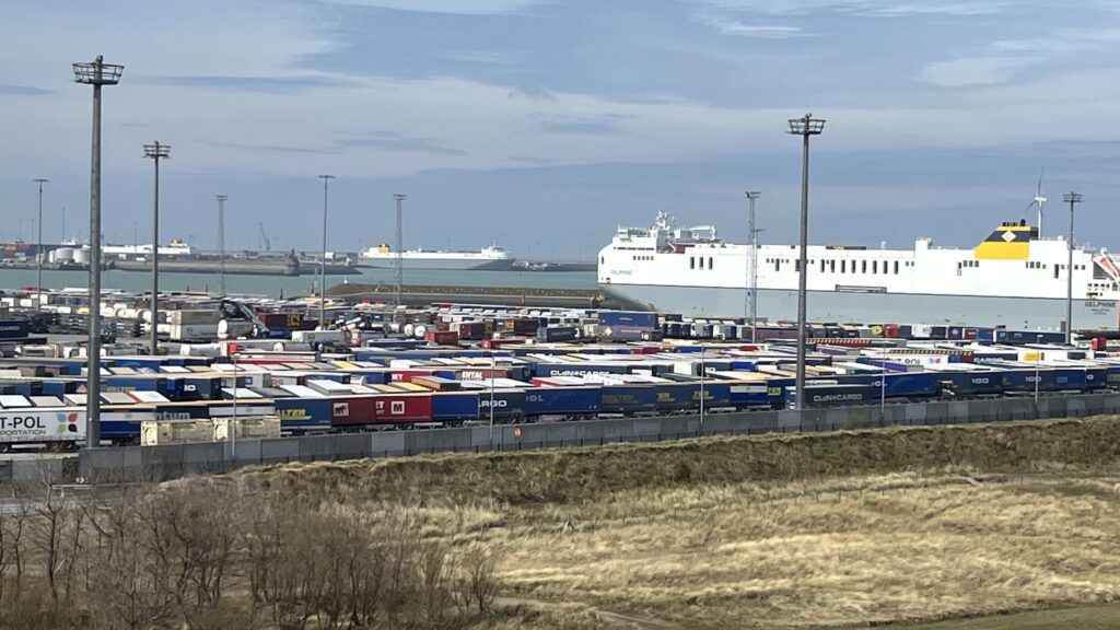 Reihen bunter LKW-Auflieger auf Hafengelände, dahinter großer weißer Frachtfährer im Wasser unter blauem Himmel.