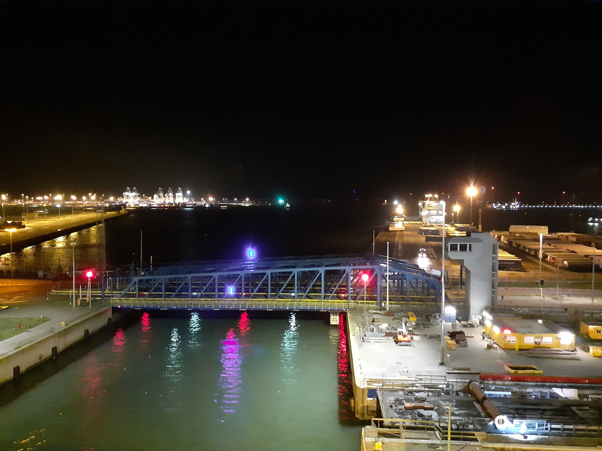 Nachthafen mit beleuchtetem blauen Anleger, rotem Signallicht, reflektierendem Wasser und erleuchteten Kränen im Hintergrund.