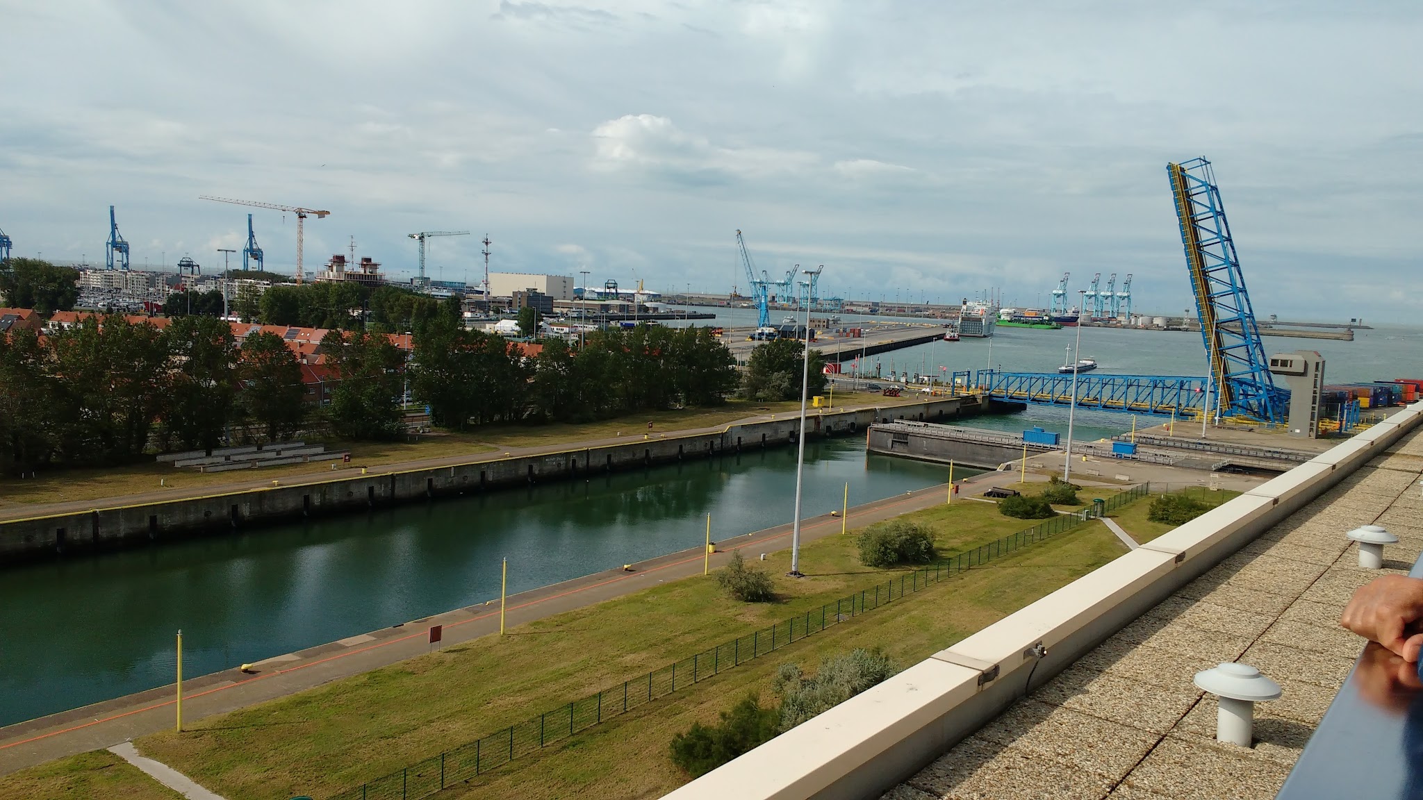 Geöffnete blaue Klappbrücke an Hafenbecken mit Schleuse, Kränen und Schiffen unter bewölktem Himmel.
