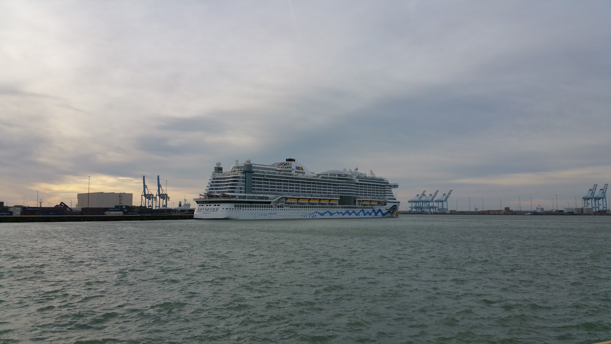 Großes Kreuzfahrtschiff liegt im Industriehafen; Containerkräne und grauer, bewölkter Himmel im Hintergrund.