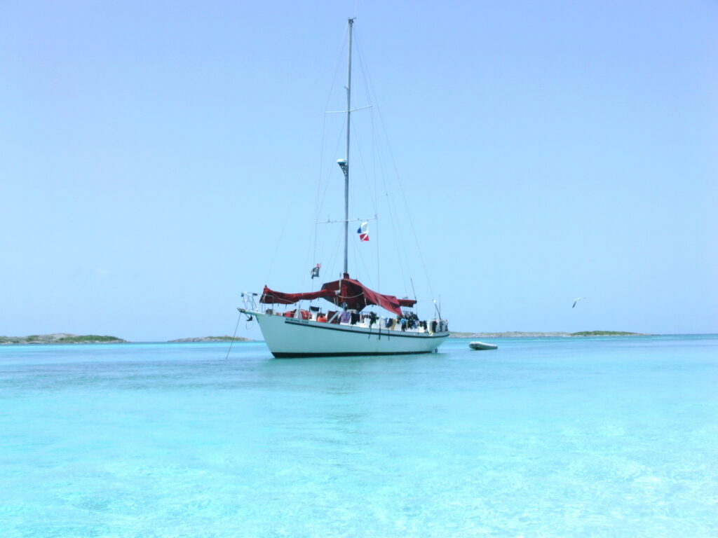 Segelboot mit rotem Sonnensegel ankert im türkisblauen, flachen Meer vor niedrigen Inseln unter klarem Himmel.