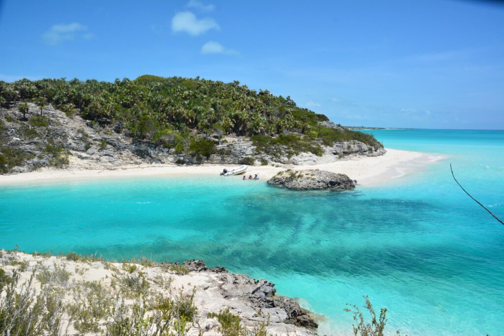 Türkisfarbenes Wasser, einsame Sandbucht mit Schlauchboot und drei Menschen, bewaldete Felsen unter blauem Himmel