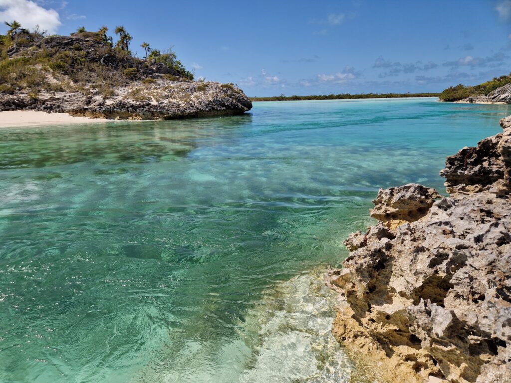 Türkisfarbene Lagune mit klarem Wasser, hellem Sandstrand links und zerklüfteten Felsen vorn unter blauem Himmel