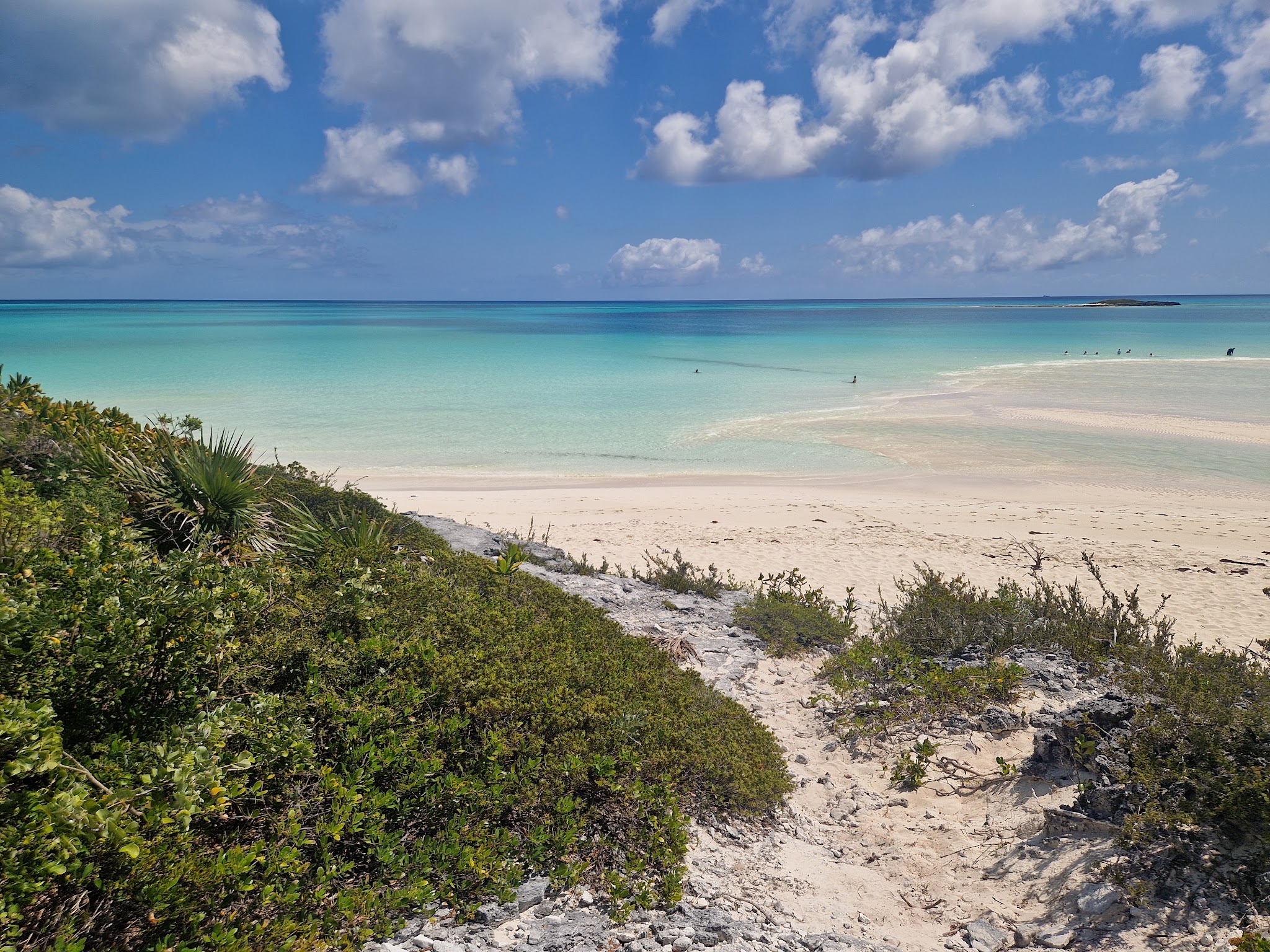 Pfad durch grüne Dünen führt zu weißem Sandstrand, türkisfarbenem Meer und blauem Himmel mit lockeren Wolken.