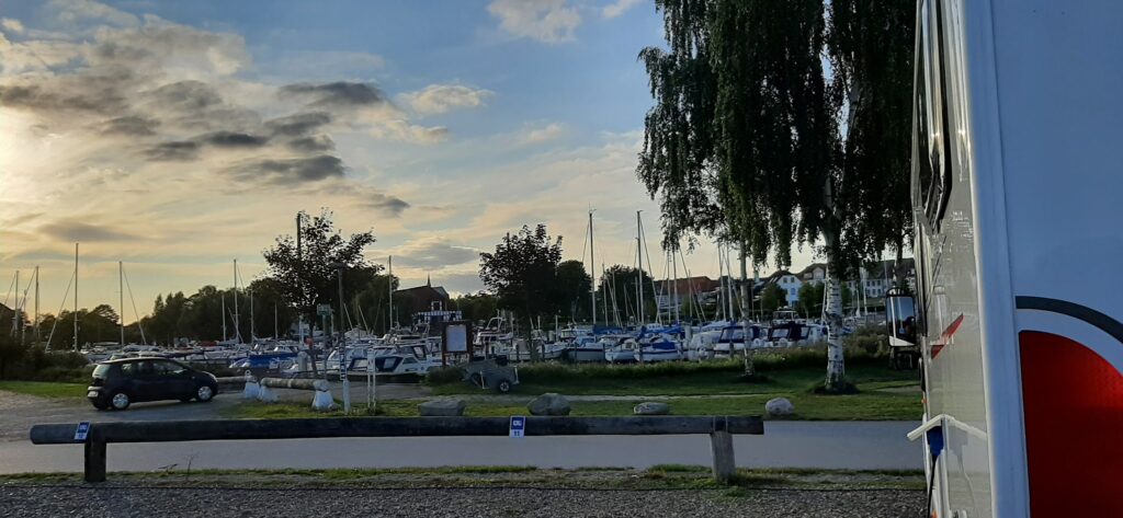 Abendstimmung am Yachthafen: zahlreiche Boote und Masten hinter Bäumen, vorne geparktes Auto, rechts ein Wohnmobil.