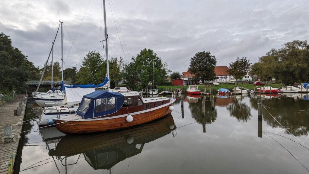 Kleiner Hafen: Holzsegelboot und weitere Boote liegen im ruhigen Wasser, Bäume und Fachwerkhaus im Hintergrund.