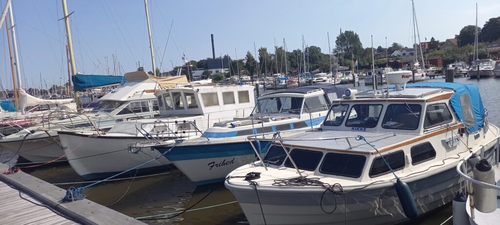Mehrere Motor- und Segelboote liegen bei Sonnenschein dicht an einem Steg im ruhigen Hafen.