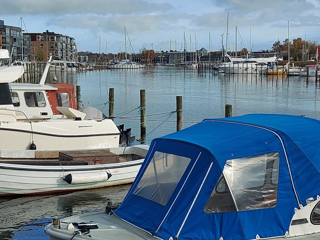 Vordergrund: kleines Motorboot mit blauem Verdeck; dahinter Hafenbecken mit Segelbooten, Pfählen und Uferhäusern.