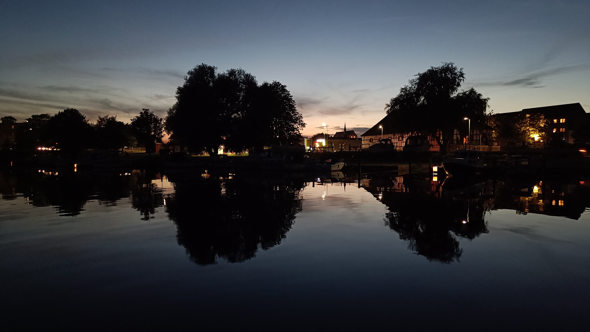 Abenddämmerung am See; dunkle Baum- und Haus-Silhouetten spiegeln sich im ruhigen Wasser mit vereinzelten Lichtern.