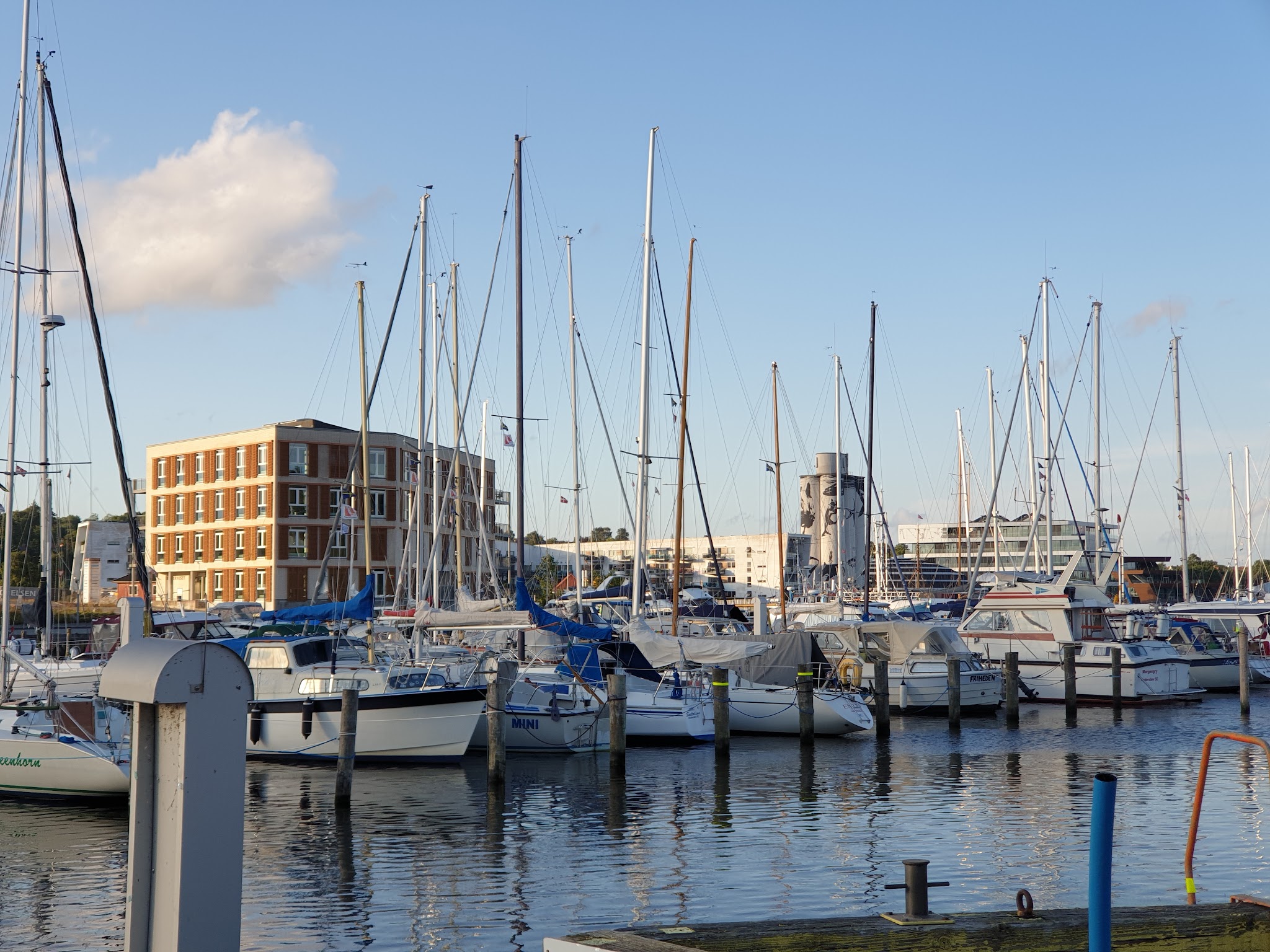 Zahlreiche Segelboote mit hohen Masten liegen in einem Yachthafen vor modernem Backsteingebäude.