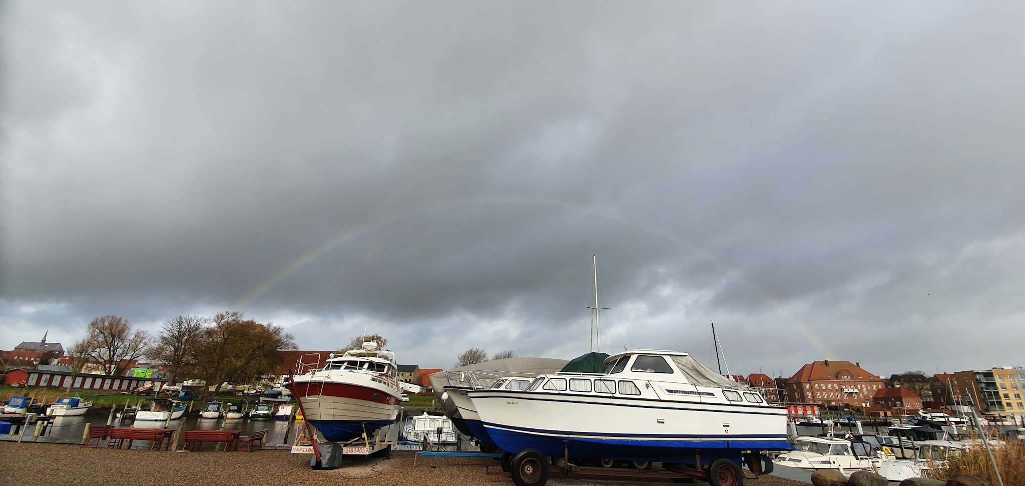Zwei aufgebockte Motorboote am Hafen, dunkle Wolken darüber, schwacher Regenbogen spannt sich über die Szene.