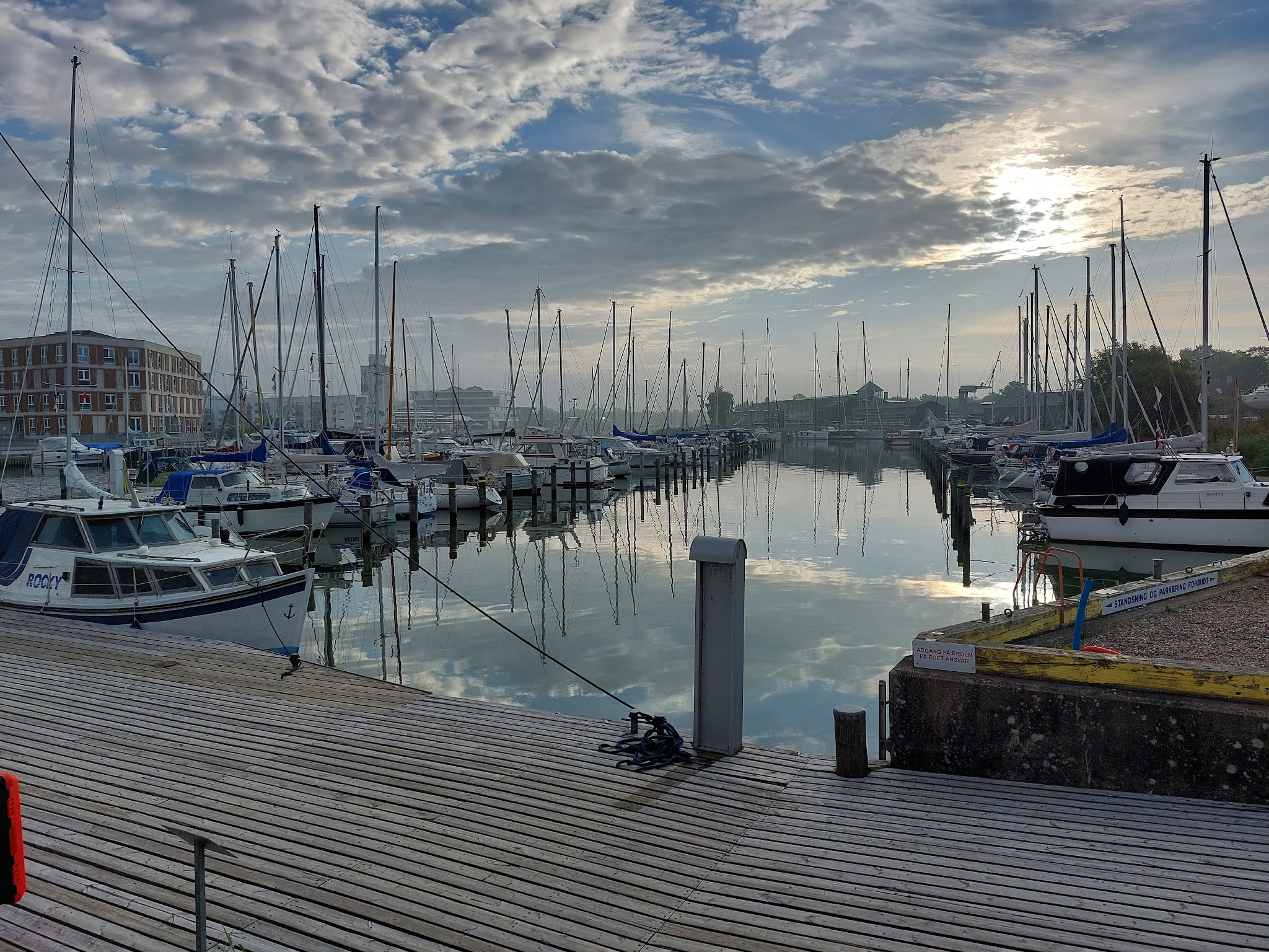 Ruhiger Yachthafen bei Morgendämmerung, vertäute Segelboote spiegeln sich im stillen Wasser unter bewölktem Himmel.