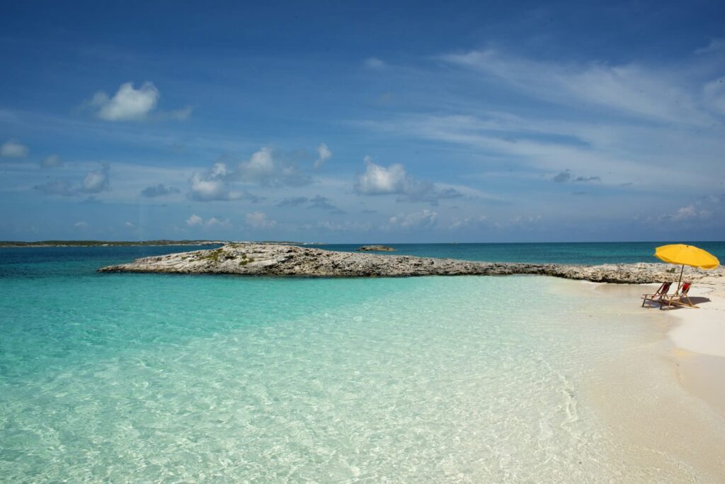 Türkisblaues, klares Meer, weißer Sand; zwei Liegestühle unter gelbem Sonnenschirm, ruhiger Felsvorsprung im Hintergrund