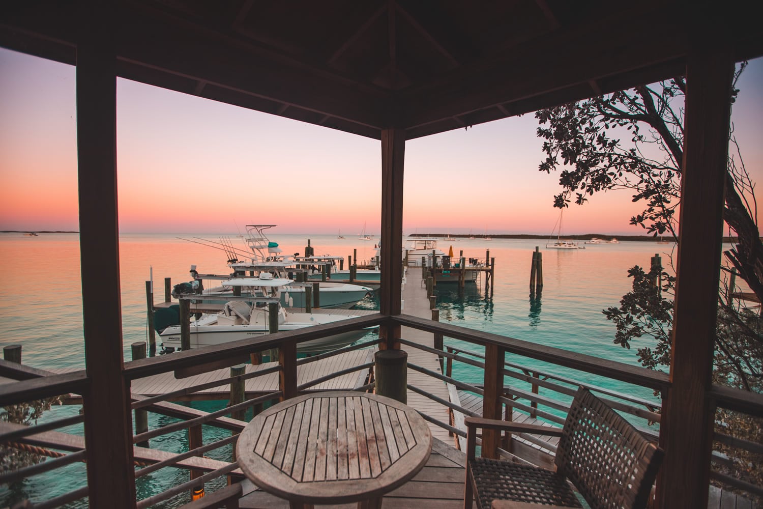 Holzterrasse mit Rundtisch, Blick auf Anleger mit Booten im türkisfarbenen Wasser bei rosa Abenddämmerung.