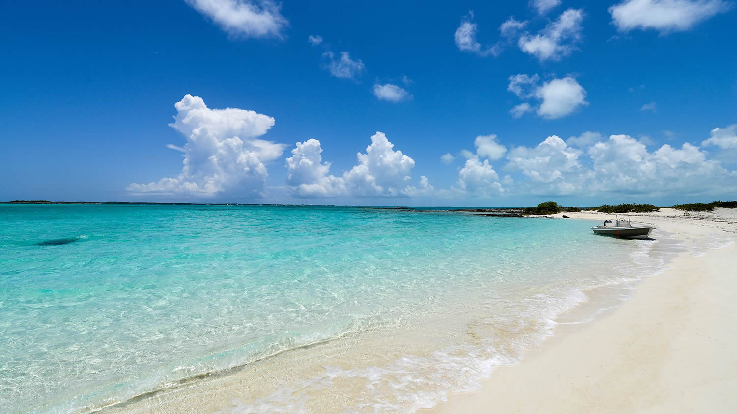 Türkisblaues, klares Meer, weißer Sandstrand, kleines Motorboot am Ufer, darüber strahlend blauer Himmel mit Quellwolken.