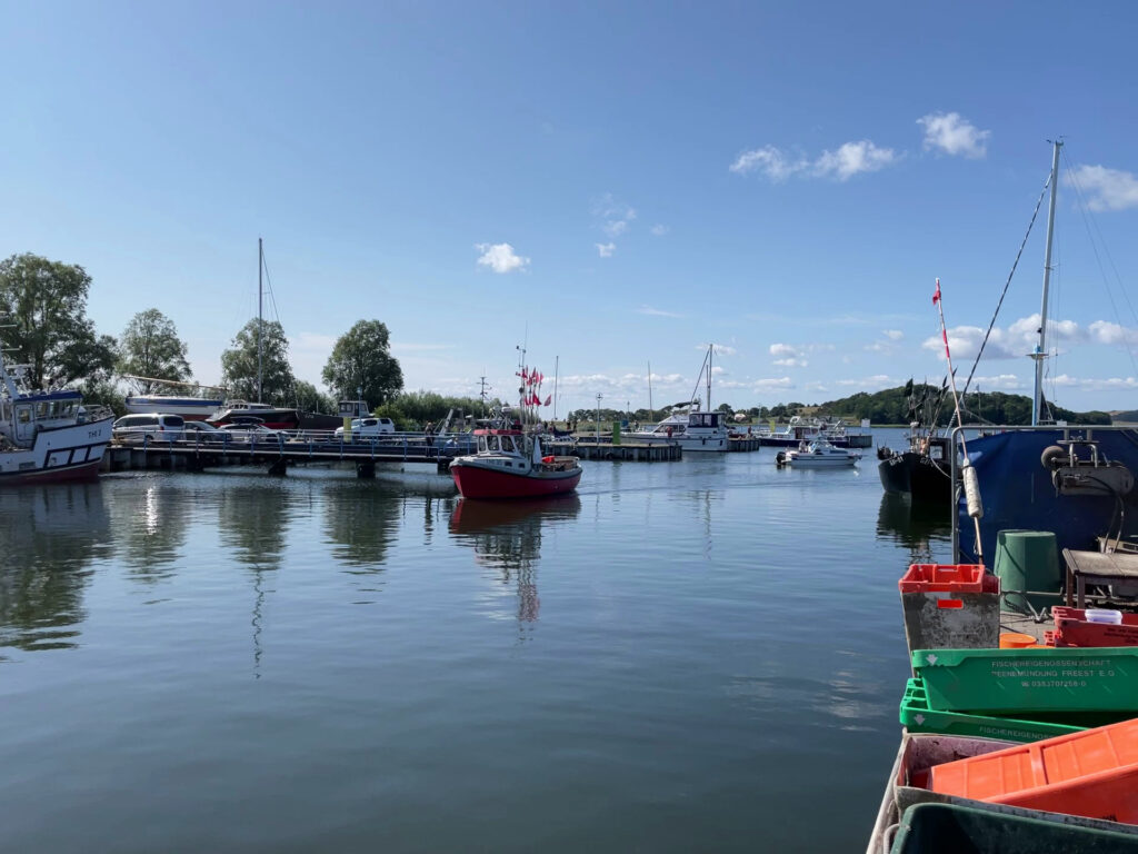 Ruhiger Hafen, mehrere Boote; rotes Fischerboot mittig, klarer blauer Himmel spiegelt sich im Wasser.