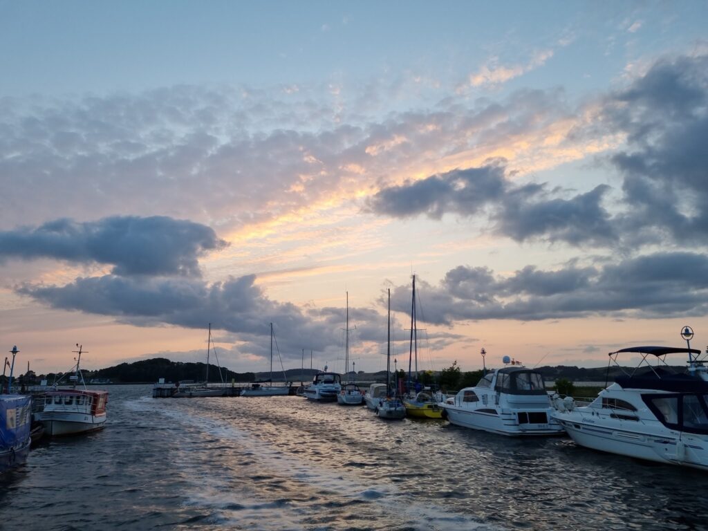 Abendhimmel über Hafen; Boote liegen am Steg, Wasser bewegt, Wolken leuchten rosa-blau.