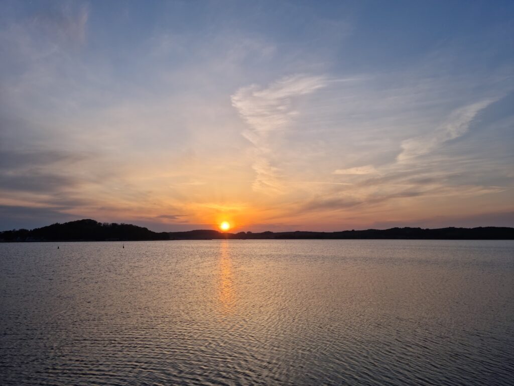 Ruhiger See bei Sonnenuntergang; orange Sonne hinter dunklen Hügeln, Himmel in Pastellblau und ‑rosa gespiegelt im Wasser
