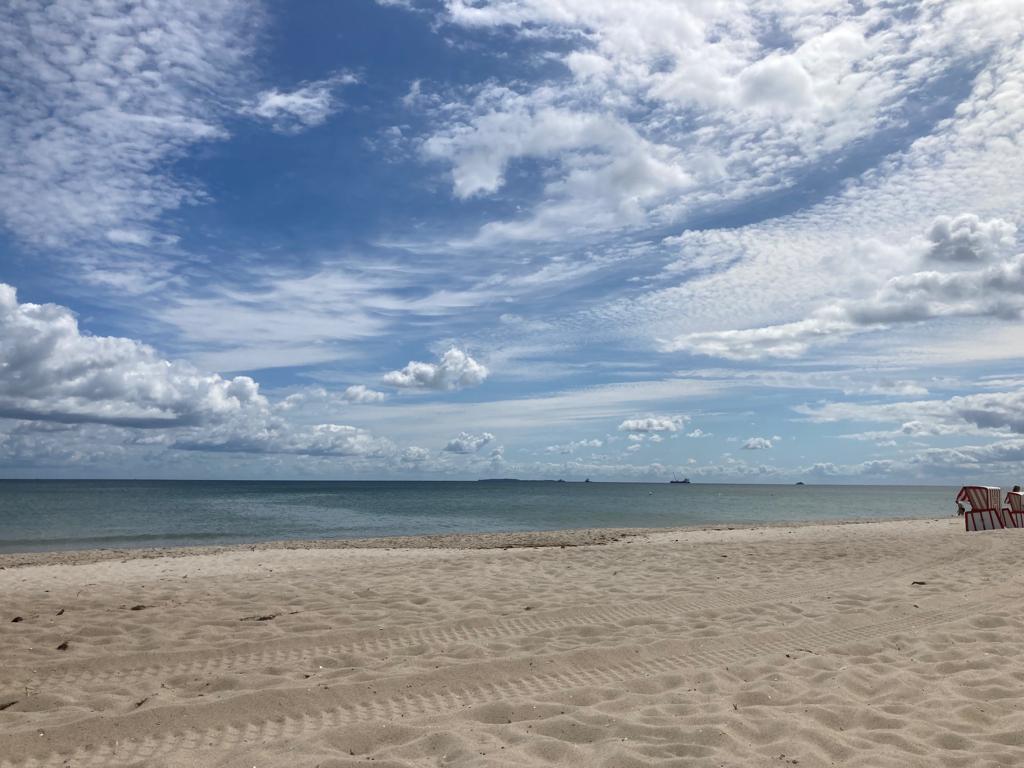 Weitläufiger Sandstrand unter teils bewölktem Himmel, ruhige See am Horizont, zwei rot-weiß gestreifte Strandstühle rechts