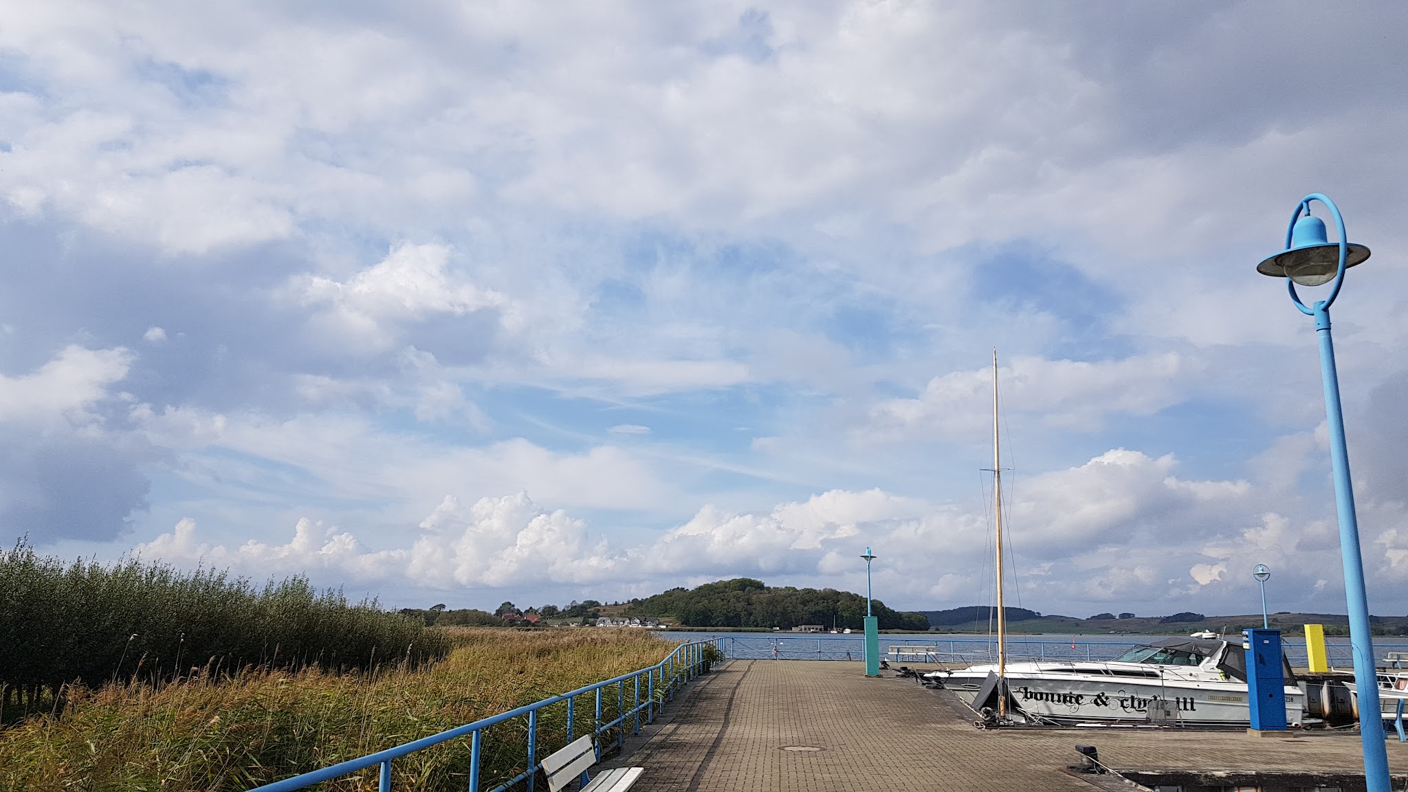 Steg mit blauem Geländer führt zwischen Schilf und festgemachten Booten auf See; bewölkter Himmel, Insel im Hintergrund