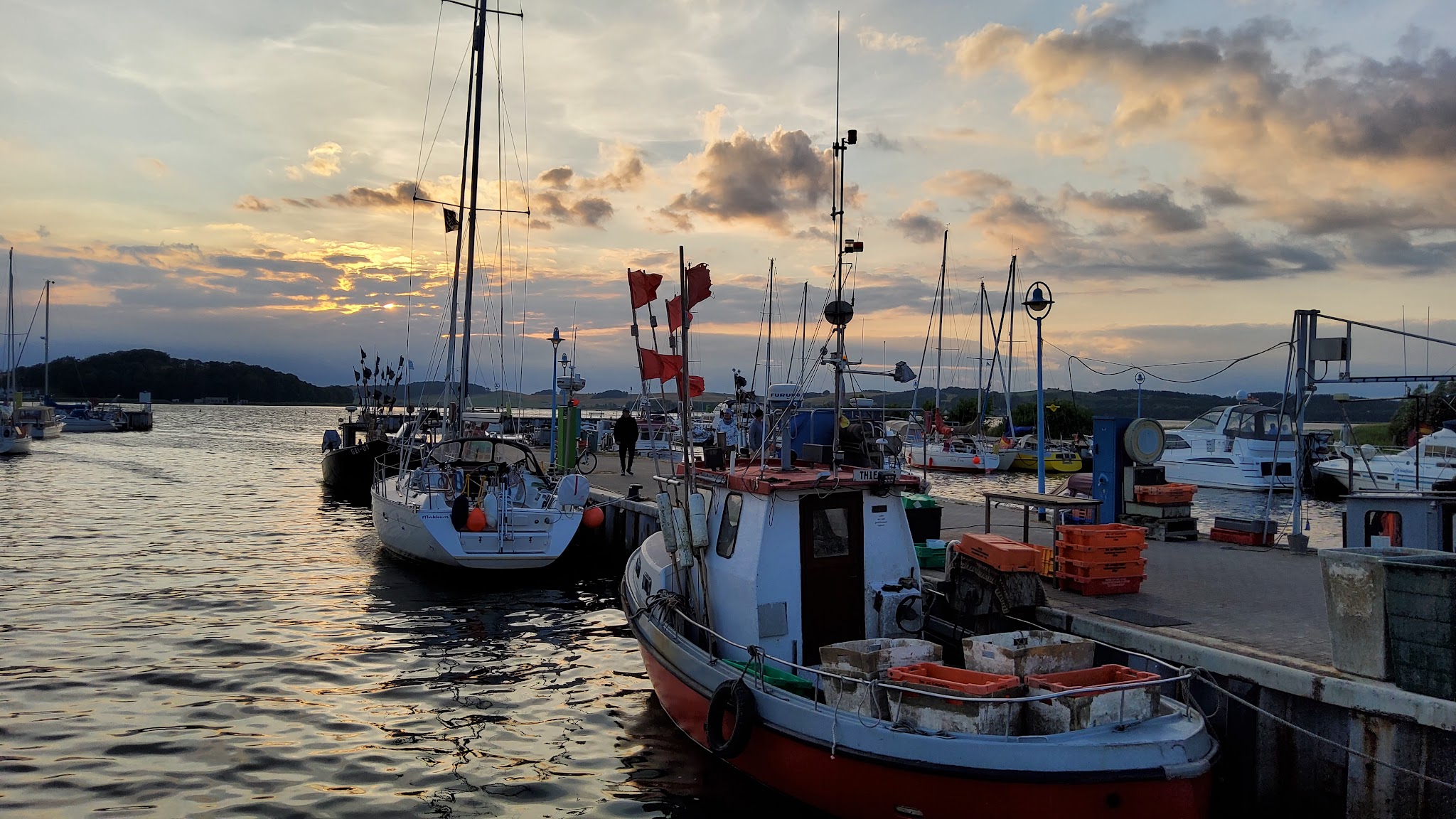 Fischerboot mit roten Flaggen und Kisten am Kai, Segelboote dahinter, Abendsonne über ruhigem Hafenwasser.