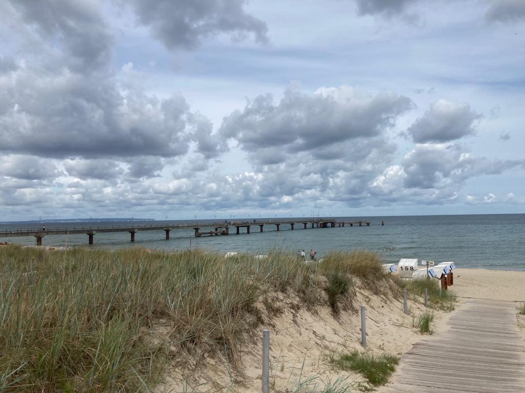 Dünengesäumter Holzsteg führt zum Sandstrand mit Strandkörben; langer Steg ragt ins ruhige Meer unter wolkigem Himmel.