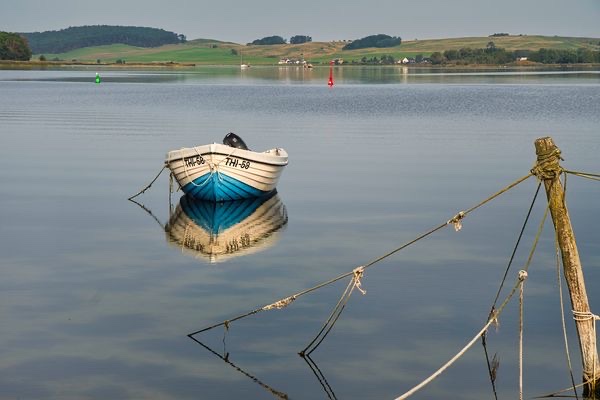 Weiß-blaues Ruderboot spiegelt sich auf ruhigem See vor sanften Hügeln; abgenutzte Seile im Vordergrund.