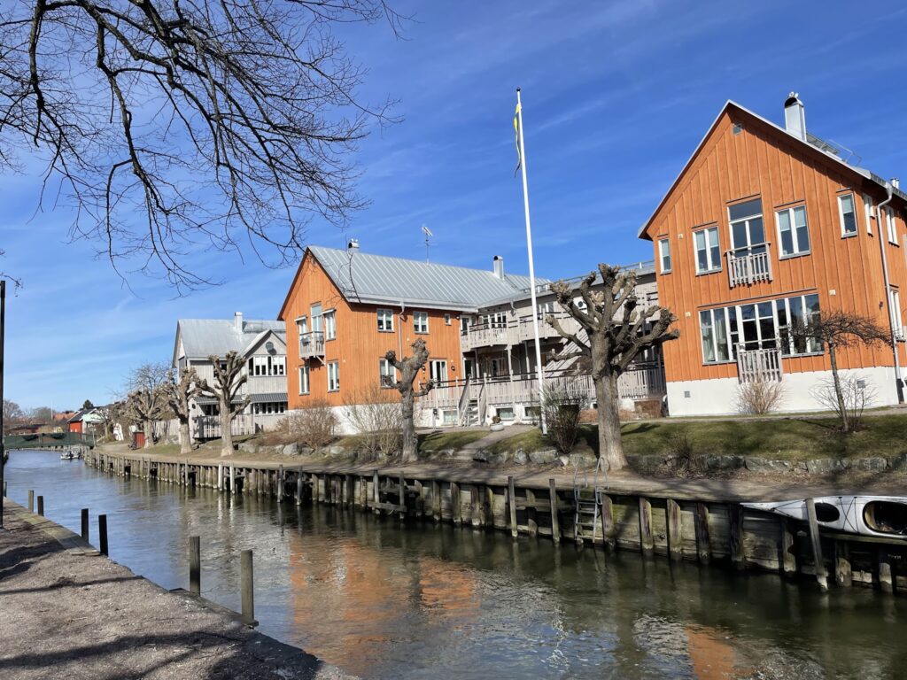 Kanalufer mit orangefarbenen Holzhäusern, kahlen Bäumen, gespiegelt im Wasser unter blauem Himmel