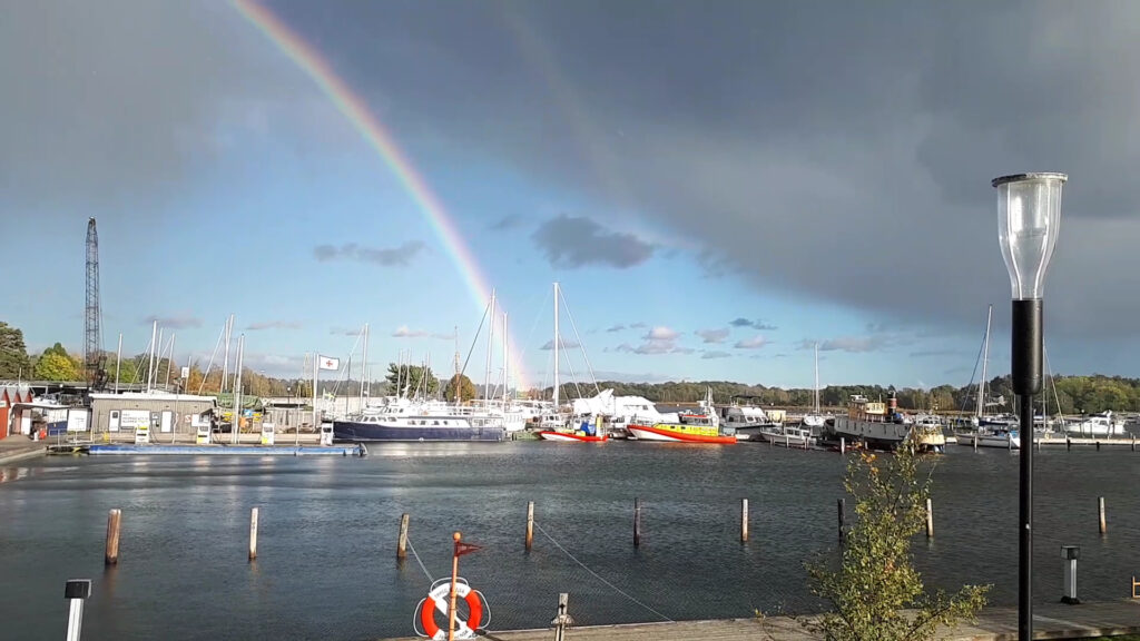 Hafen mit Segelbooten; über dem Wasser spannt sich ein Doppelregenbogen vor dunklen Gewitterwolken