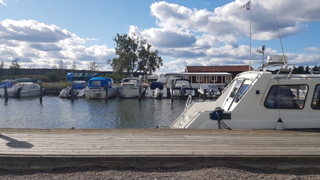 Kleine Marina; links Reihe kleiner Boote an Holzstegen, rechts Bug eines großen Kajütboots, ruhiges Wasser, Wolkenhimmel.