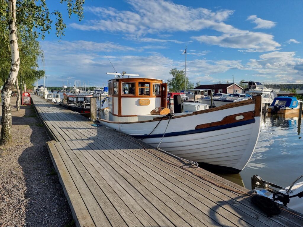 Weißes Holzboot liegt an holzbeplanktem Steg in sonnigem Yachthafen, ruhiges Wasser und blauer Himmel