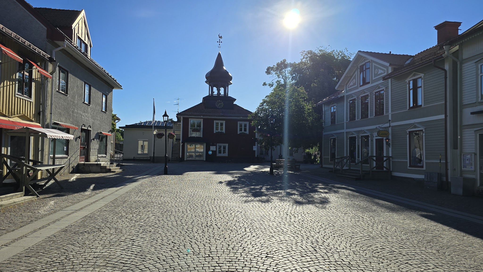 Leere Kopfsteinpflaster-Gasse, flankiert von Holzhäusern; in der Mitte rotes Rathaus mit Turmuhr unter strahlender Sonne.