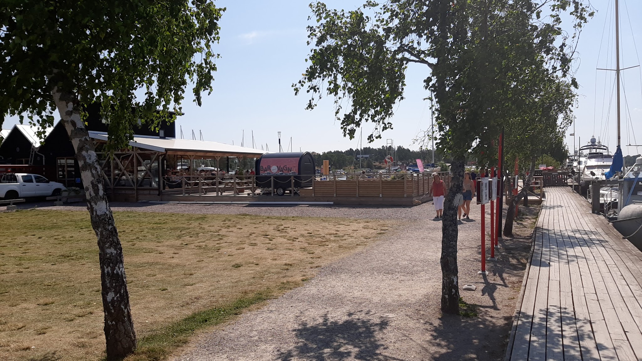 Hafencafé mit Sitzbänken unter Dach, Spazierweg am Steg, Bäume spenden Schatten, Boote im Hintergrund.