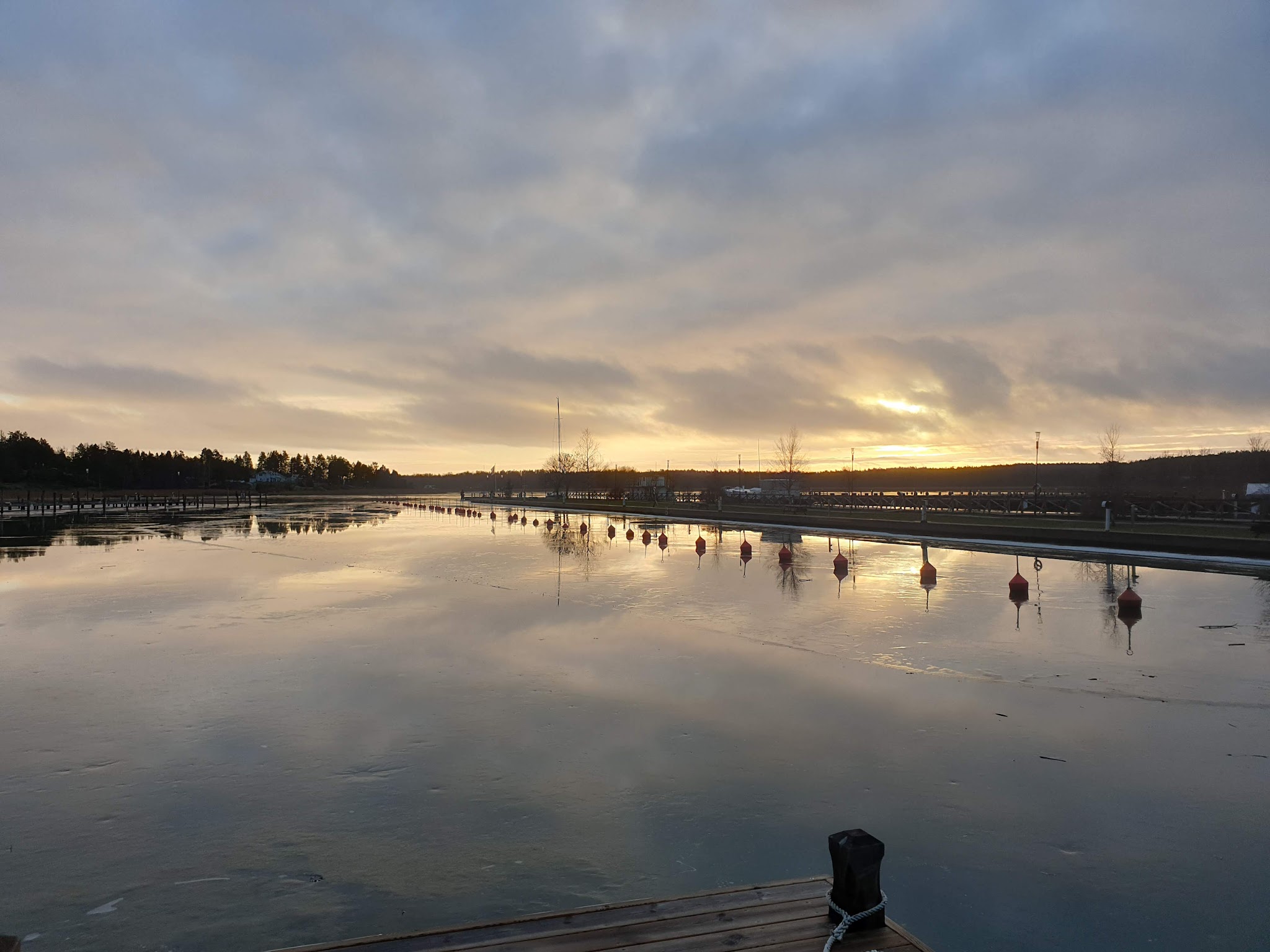 Winterlicher Hafen bei Sonnenuntergang; ruhiges Wasser spiegelt Wolken, rote Bojen markieren zwei Reihen.