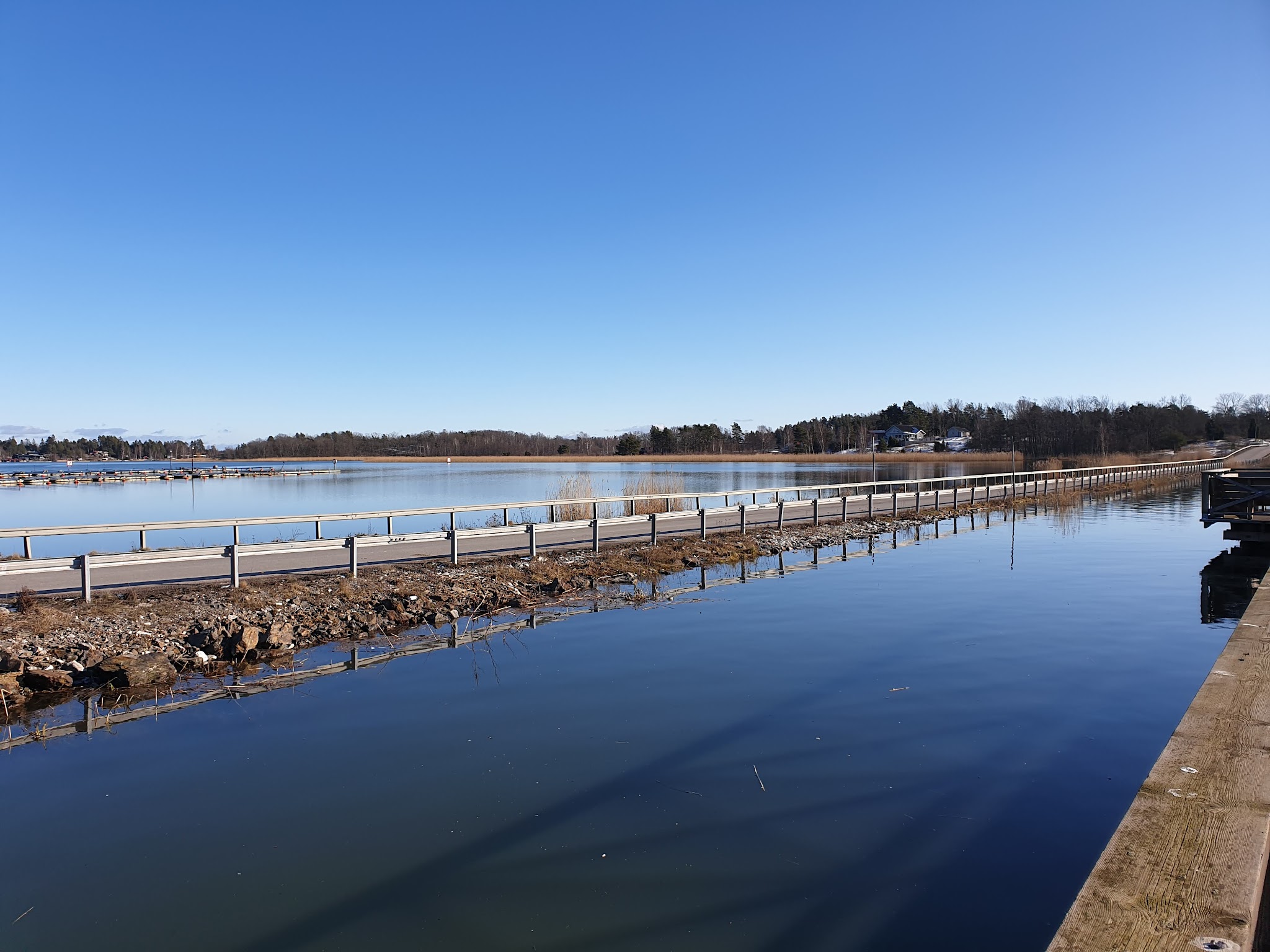 Schmaler Deichweg trennt ruhigen See und Kanal unter klarem blauem Himmel; Ufer mit Schilf und wenigen Häusern.
