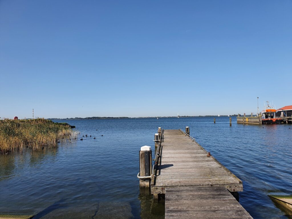 Holzsteg führt ins ruhige blaue Wasser; links Schilf mit Enten, rechts kleines Boot, ferne Segel am Horizont