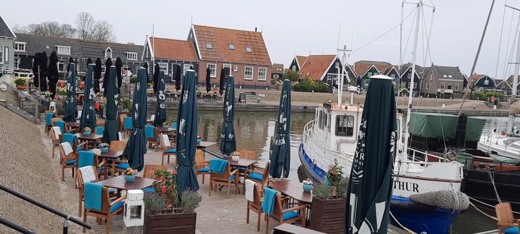 Leere Hafenterrasse mit Holztischen, geschlossenen dunklen Sonnenschirmen und Blaustühlen; Boot am Kai, Giebelhäuser dahinter