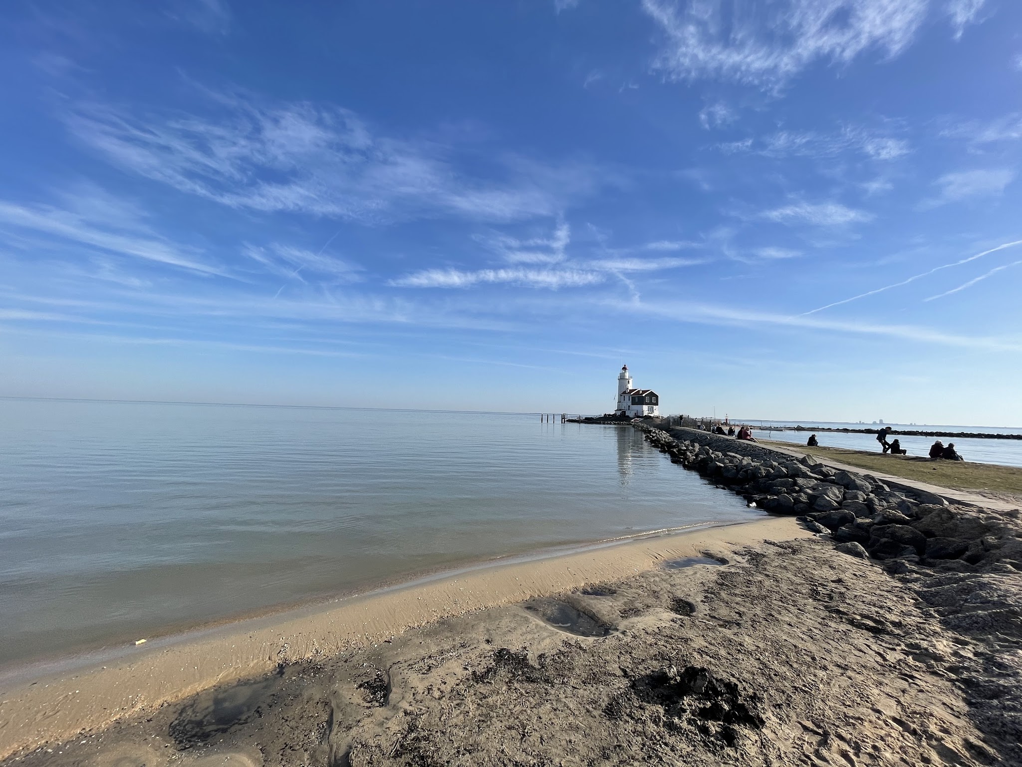 Ruhiger Strand mit Steindamm zu kleinem weißem Leuchtturm; wenige Menschen genießen Sonne unter blauem Himmel