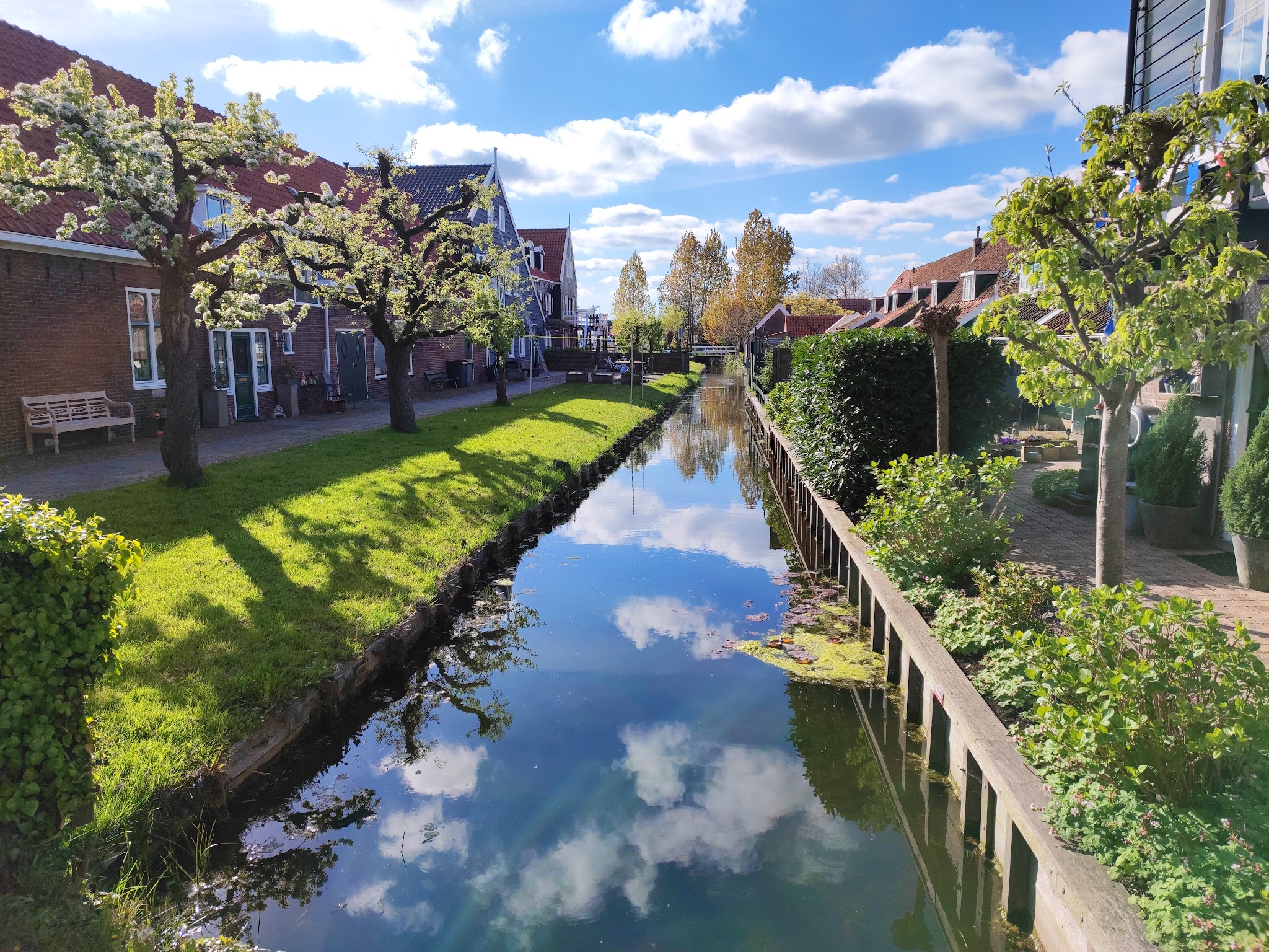 Schmaler Kanal zwischen Reihenhäusern; blühende Bäume, grüner Rasen, blau-weißer Himmel spiegelt sich im Wasser.