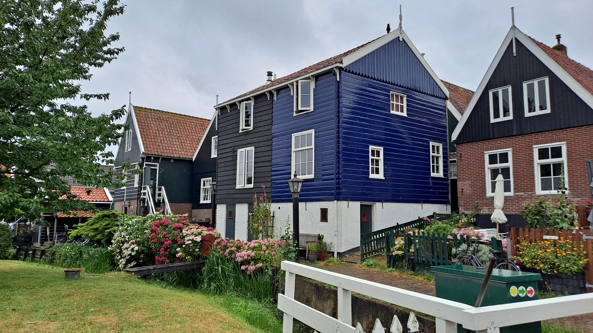 Bunte niederländische Giebelhäuser am Kanal, umgeben von üppigen Blumenbeeten und weißem Gartenzaun.