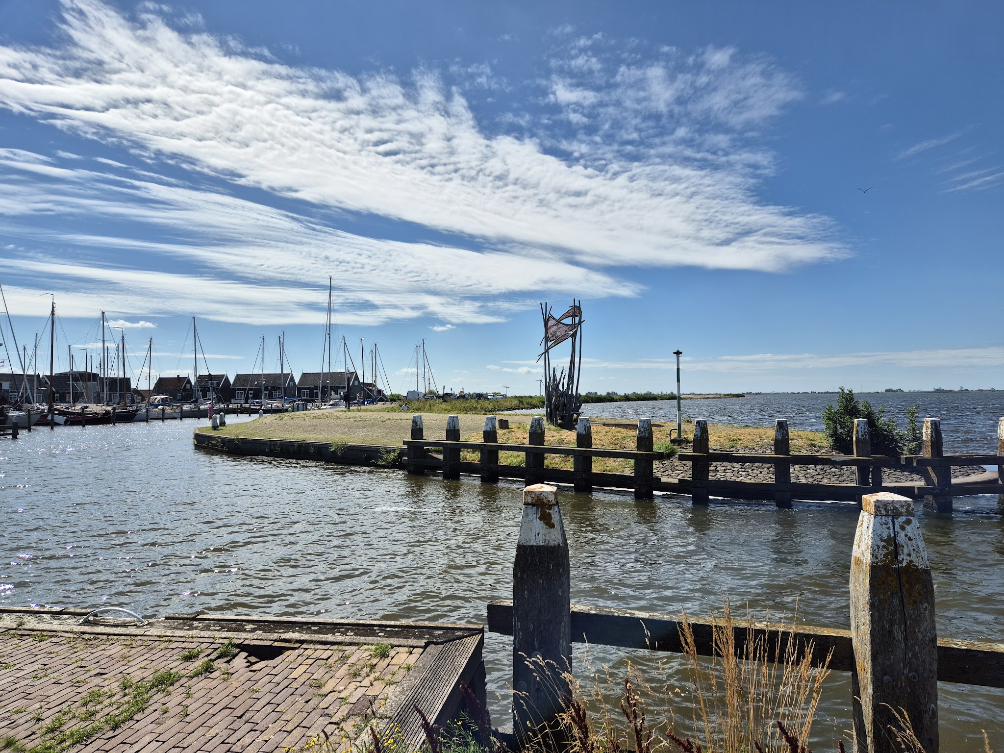 Kleiner Hafen mit vertäuten Segelbooten, Holzpfählen und Skulptur auf Landzunge unter blauem Himmel mit Wolken.