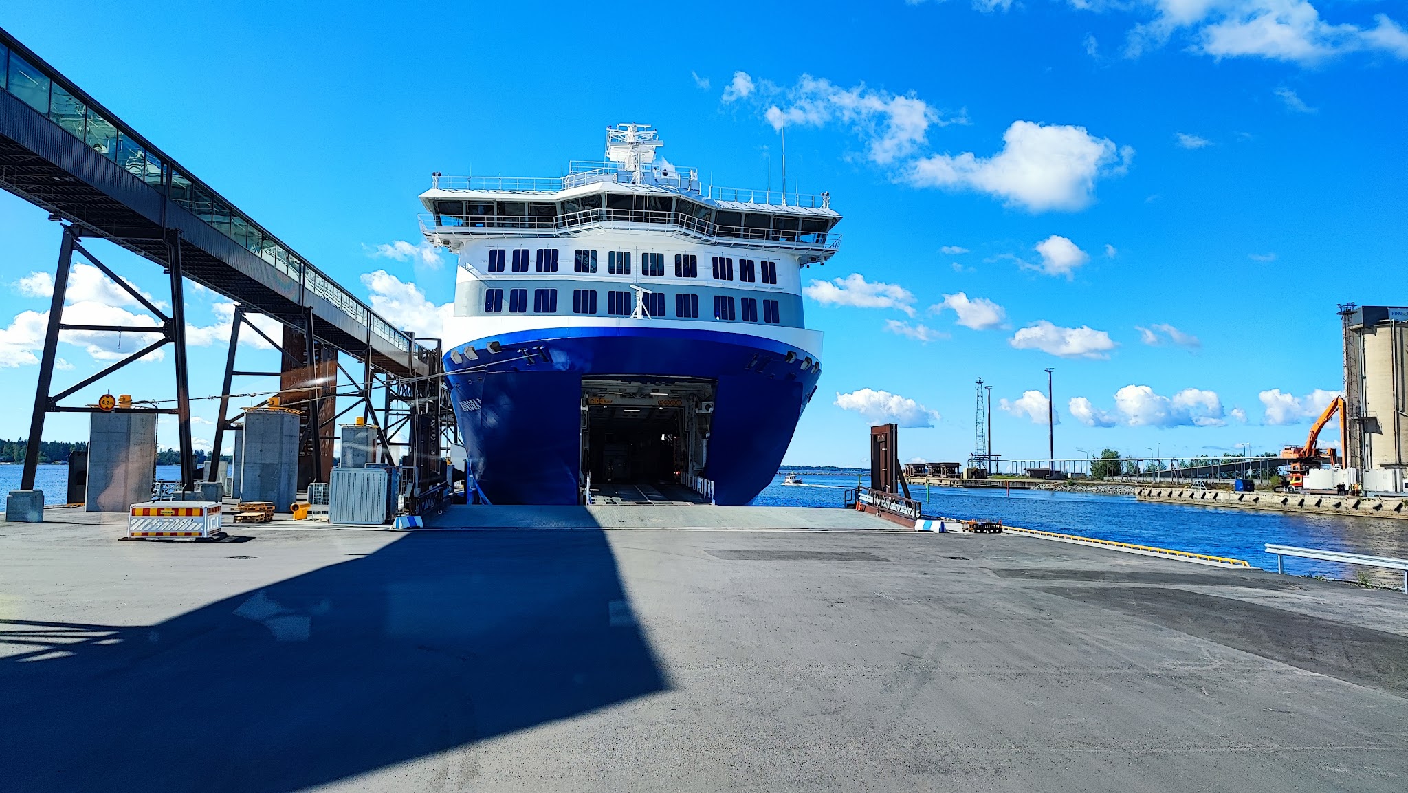 Große blau-weiße Fähre liegt am Kai, Bugrampe offen; links verglaster Fußgängersteg, klarer Himmel über dem Hafen