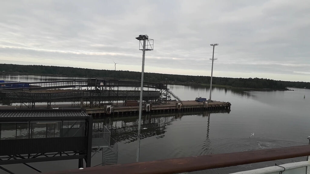 Hafensteg mit Metallgängen und hohen Lampenmasten, ruhiges Wasser vor Waldufer, grauer Himmel, Windrad.