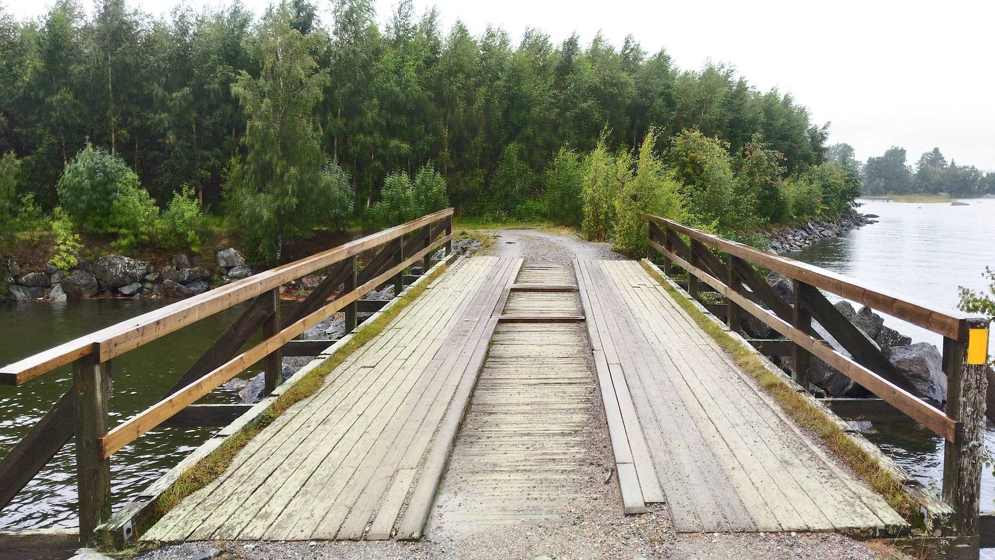 Schmale Holzbrücke mit Geländern führt über ruhiges Wasser zu einem Waldweg.